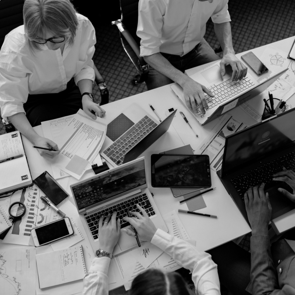 A team working across laptops, printed charts, and marked-up documents around one table, seen from above
