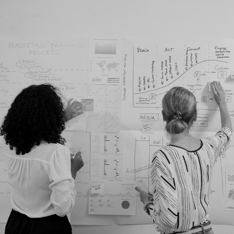 Two colleagues mapping a process across a wall of printed charts and hand-drawn diagrams